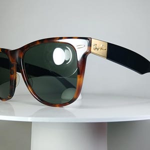 May include: Tortoiseshell-framed sunglasses with dark green lenses. The sunglasses have a gold accent on the temple and black arms. The Ray-Ban logo is visible on the temple. The sunglasses are displayed on a white surface.