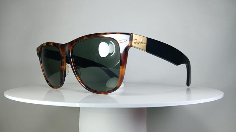 May include: Tortoiseshell-framed sunglasses with dark green lenses. The sunglasses have a gold accent on the temple and black arms. The Ray-Ban logo is visible on the temple. The sunglasses are displayed on a white surface.