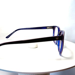May include: Blue and black eyeglasses with a rectangular frame. The glasses have a dark blue frame with a black arm and a small silver emblem. The glasses are displayed on a white surface.