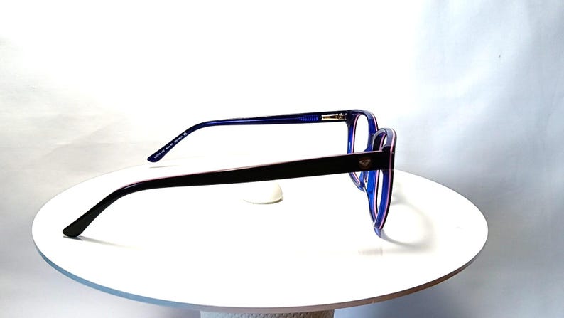May include: Blue and black eyeglasses with a rectangular frame. The glasses have a dark blue frame with a black arm and a small silver emblem. The glasses are displayed on a white surface.