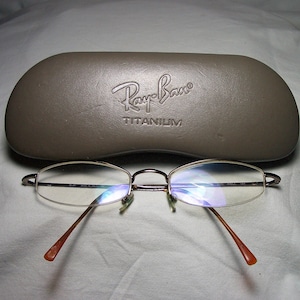 May include: A brown leather Ray-Ban glasses case with the text "Ray-Ban TITANIUM" embossed on the front. A pair of silver-rimmed glasses with clear lenses are sitting on a white surface.