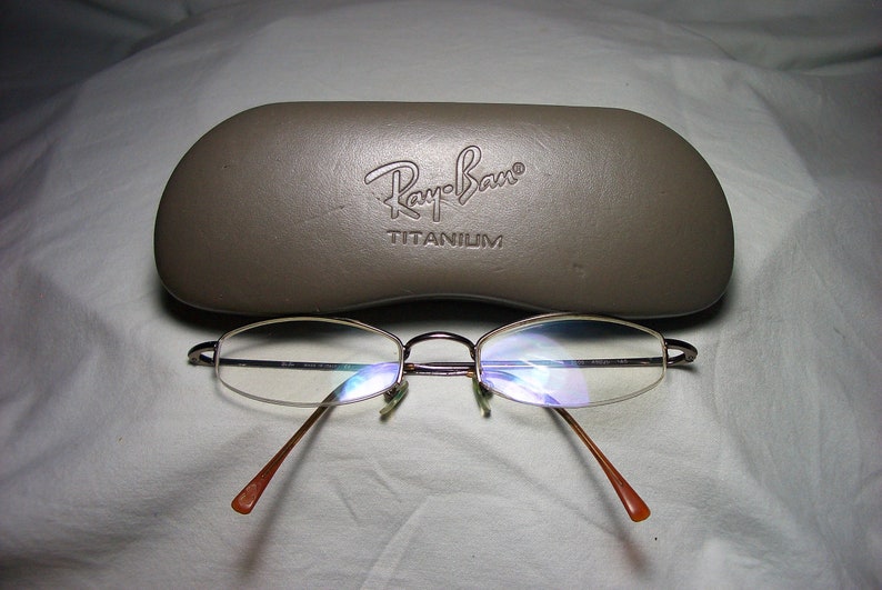 May include: A brown leather Ray-Ban glasses case with the text "Ray-Ban TITANIUM" embossed on the front. A pair of silver-rimmed glasses with clear lenses are sitting on a white surface.