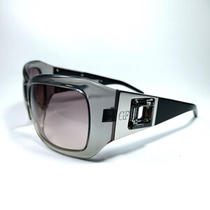 May include: Silver and black sunglasses with a large, rectangular frame and a clear lens. The sunglasses have a silver metal detail on the side with the letters "GF" embossed on them.