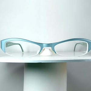Puede incluir: Un par de gafas con montura azul claro mate y un diseño angular y elevado. Las lentes son transparentes y no tienen montura en el borde inferior, con varillas finas de color azul claro a juego que se curvan hacia el interior.