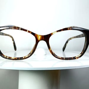 May include: Cat-eye shaped spectacles featuring a glossy brown and black tortoiseshell pattern on the frame front and temples. The glasses have a distinct upswept outer edge design and clear lenses, showcasing a classic look with a polished finish.