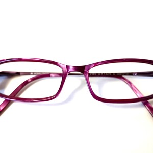 Peut inclure: Une paire de lunettes rectangulaires avec une monture violette marbrée. Les lunettes présentent une couleur violet foncé sur les bords extérieurs, qui s'estompe en un violet plus clair vers les bords intérieurs. Les branches et le pont de nez sont violets assortis.