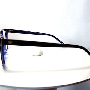 May include: Black and blue eyeglasses with a rectangular frame. The glasses have a dark frame with a small gold emblem on the side. The lenses are blue, and the arms are thin and black. The glasses are displayed on a white surface.