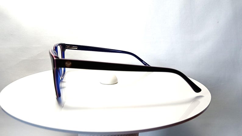 May include: Black and blue eyeglasses with a rectangular frame. The glasses have a dark frame with a small gold emblem on the side. The lenses are blue, and the arms are thin and black. The glasses are displayed on a white surface.
