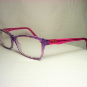 Puede incluir: Gafas de pl&aacute;stico con montura de color morado y rosa con lentes transparentes. El nombre de la marca "Ray-Ban" es visible en la patilla derecha.