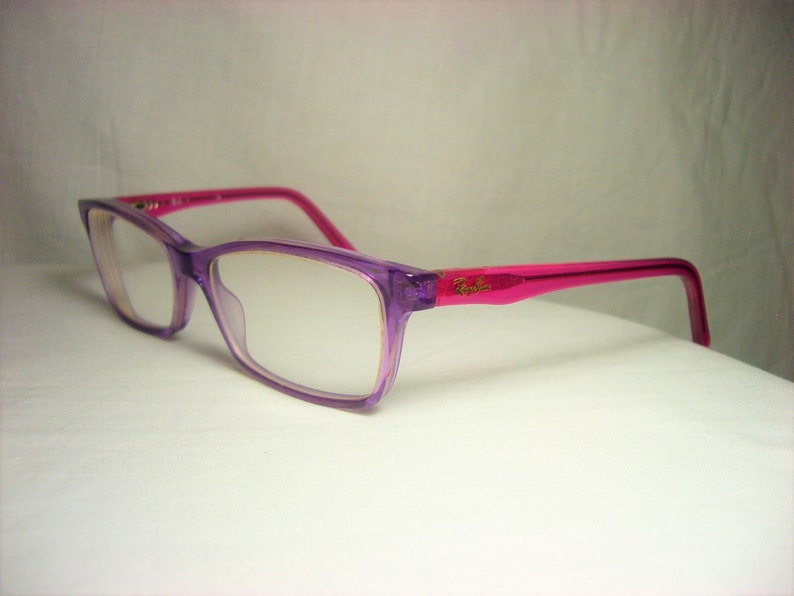 Puede incluir: Gafas de pl&aacute;stico con montura de color morado y rosa con lentes transparentes. El nombre de la marca "Ray-Ban" es visible en la patilla derecha.