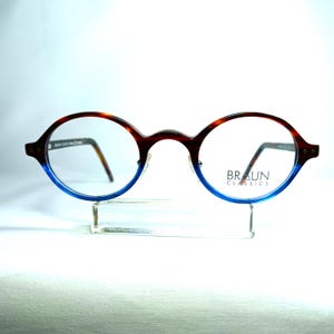 May include: Oval-shaped eyeglasses with a brown and blue gradient frame. The top of the frame is brown with a tortoise shell pattern, transitioning to blue at the bottom. The lenses are clear, and the brand name "BRAUN CLASSICS" is printed on the right lens.