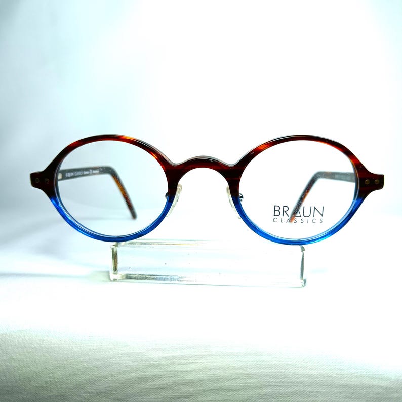 May include: Oval-shaped eyeglasses with a brown and blue gradient frame. The top of the frame is brown with a tortoise shell pattern, transitioning to blue at the bottom. The lenses are clear, and the brand name "BRAUN CLASSICS" is printed on the right lens.