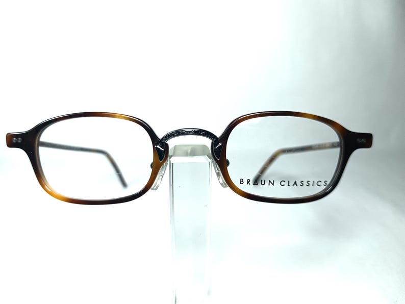 May include: Brown and black oval-shaped eyeglasses with clear lenses. The frames have a tortoiseshell pattern and a silver bridge. The brand name "BRAUN CLASSICS" is printed on the right lens.
