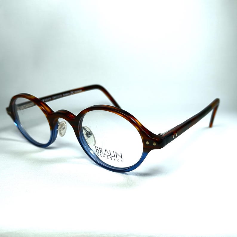 May include: Oval-shaped eyeglasses with a brown and blue tortoiseshell pattern. The frames have a blue gradient at the bottom and the brand name "BRAUN CLASSICS" is visible on the lens.