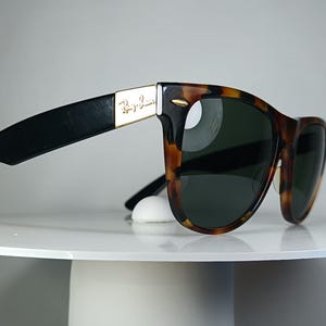 May include: Sunglasses with a tortoiseshell pattern frame and dark lenses. The temples are black with a gold accent and the Ray-Ban logo. The sunglasses are resting on a white, round surface.