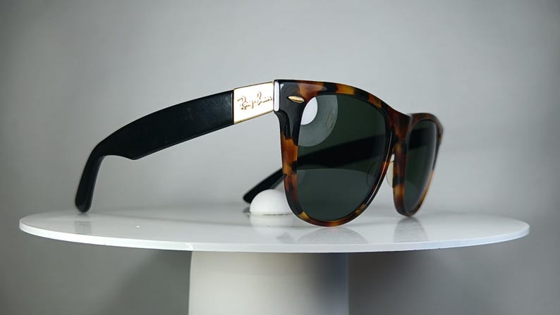 May include: Sunglasses with a tortoiseshell pattern frame and dark lenses. The temples are black with a gold accent and the Ray-Ban logo. The sunglasses are resting on a white, round surface.