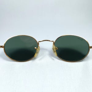 Puede incluir: Gafas de sol ovaladas con montura dorada y lentes verdes. Las gafas de sol est&aacute;n sobre un fondo blanco.