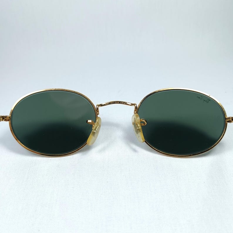 Puede incluir: Gafas de sol ovaladas con montura dorada y lentes verdes. Las gafas de sol est&aacute;n sobre un fondo blanco.