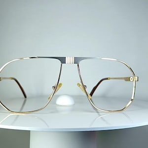 May include: Gold-framed aviator spectacles with a silver bridge and brown arms. The glasses feature a classic, oversized design and are displayed on a white surface against a neutral background.