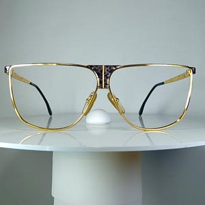 May include: Gold-rimmed eyeglasses with a square shape and black temples. The bridge features a patterned design. The glasses are displayed on a white pedestal, showcasing a vintage or retro style.
