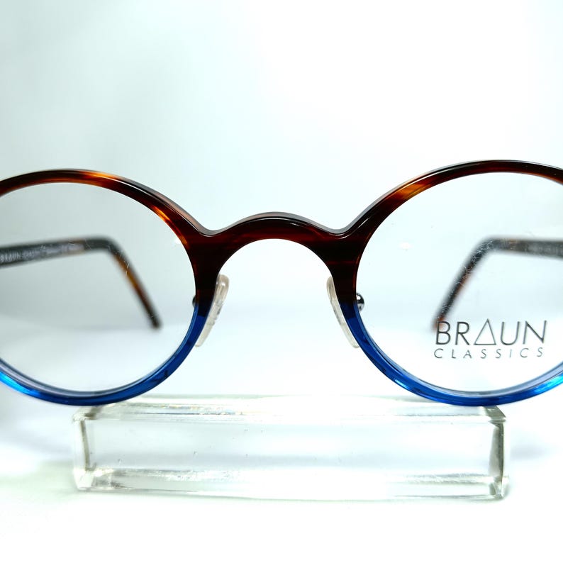 May include: Round eyeglasses with a brown and blue gradient frame. The top of the frame is brown with a tortoise shell pattern, transitioning to blue at the bottom. The brand name "BRAUN CLASSICS" is printed on the right lens.