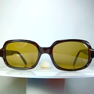 May include: A pair of sunglasses featuring a dark brown, thick-rimmed plastic frame with a rounded rectangular shape. The lenses are a solid, translucent amber-brown color, providing a warm tint. The temples are visible extending from the sides.