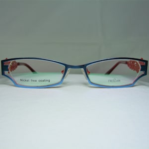 May include: Blue metal eyeglasses with red accents and a nickel-free coating. The lenses are clear and the brand name "FRESCURA" is printed on the right lens.