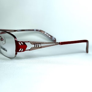 Op de afbeelding: Rode en zilveren bril met een rechthoekig montuur. De veren hebben een decoratief patroon en de merknaam "D'Addio" is zichtbaar op de lens. De bril heeft een modern design.