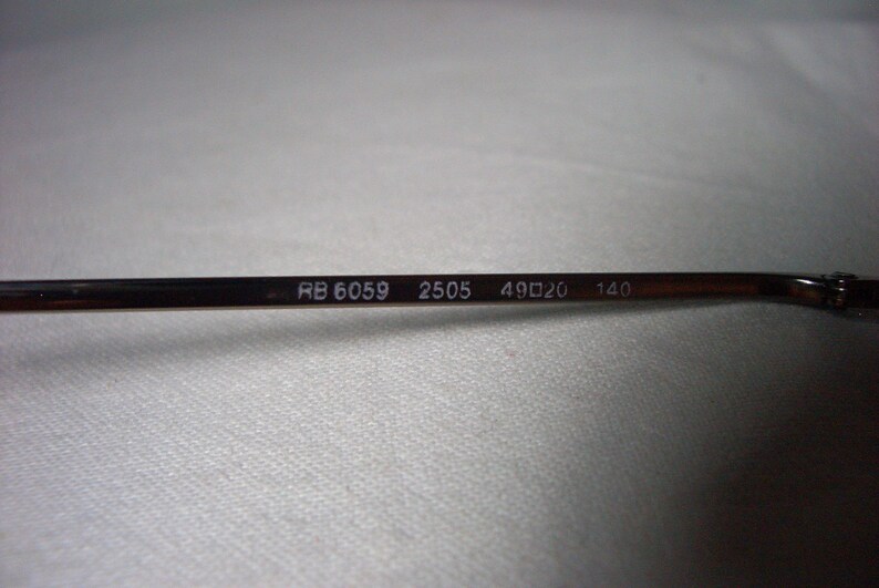May include: Close-up of the inside of a pair of glasses with the text "RB 6059 2505 49□20 140" printed on the metal arm.