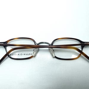 May include: Eyeglasses with a brown tortoiseshell pattern on the frames and temples. The glasses have a silver bridge and clear lenses. The brand name "BRAUN CLASSICS" is printed on the inside of the frame.