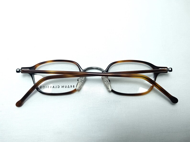 May include: Eyeglasses with a brown tortoiseshell pattern on the frames and temples. The glasses have a silver bridge and clear lenses. The brand name "BRAUN CLASSICS" is printed on the inside of the frame.