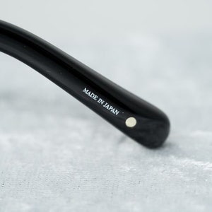 Puede incluir: Primer plano de una montura de gafas negra con el texto "MADE IN JAPAN" impreso en el lateral.