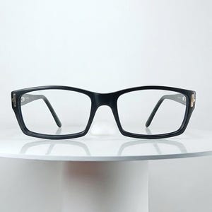以下が含まれることがあります： 外側のヒンジにT字型の金属製ディテールが施された、厚みのある黒い長方形のメガネ。透明なレンズは、ブリッジでつながった頑丈な黒いプラスチックフレームに収められており、すっきりとしたデザインが特徴です。