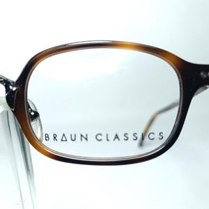 May include: Close-up of eyeglasses with a brown tortoiseshell frame. The glasses have a classic, rounded shape and clear lenses. The text "BRAUN CLASSICS" is printed on the lens. The frame is a mix of light and dark brown.