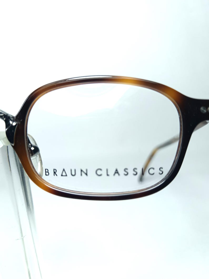 May include: Close-up of eyeglasses with a brown tortoiseshell frame. The glasses have a classic, rounded shape and clear lenses. The text "BRAUN CLASSICS" is printed on the lens. The frame is a mix of light and dark brown.