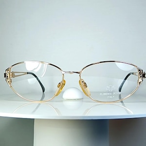 Könnte beinhalten: Goldfarbene Brille mit ovalen Gläsern und schwarzen Bügeln. Die Fassungen haben dekorative Akzente an den Seiten und einen Steg mit Nasenpads. Die Brille wird auf einer weißen, runden Oberfläche präsentiert.