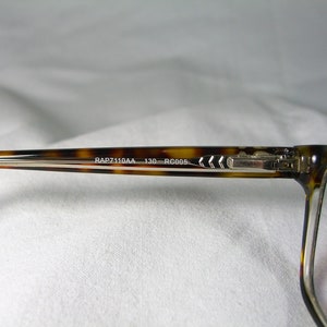 May include: Tortoise shell eyeglasses with silver metal arms and a clear lens. The arms are engraved with the text "RAP7110AA 130 RC005."