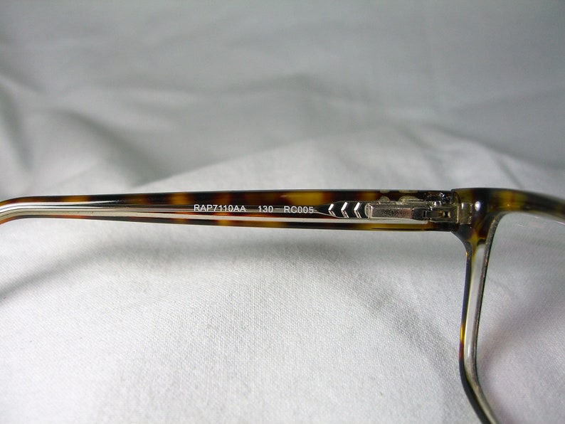 May include: Tortoise shell eyeglasses with silver metal arms and a clear lens. The arms are engraved with the text "RAP7110AA 130 RC005."