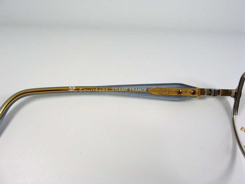 May include: A close-up of the arm of a pair of eyeglasses. The arm is made of a light blue metal with a gold trim. The text "JL COUTURIER FRAME FRANCE" is engraved on the arm.