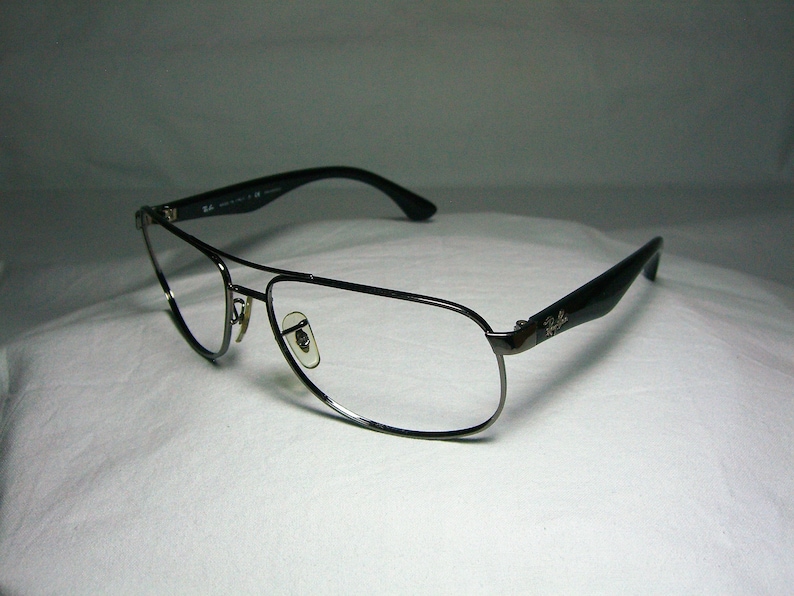 K&ouml;nnte beinhalten: Eine silberfarbene Aviator-Brille mit schwarzen B&uuml;geln. Die Brille hat klare Gl&auml;ser und ein Ray-Ban-Logo am B&uuml;gel.