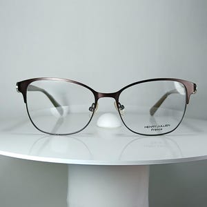 Könnte beinhalten: Dunkelbraune Metallbrille mit katzenaugenförmigem Rahmen. Die Gläser sind klar und auf dem rechten Glas befindet sich der Aufdruck HENRY JULLIEN France. Die Brille verfügt über dünne Bügel und verstellbare Nasenpads für einen eleganten Look.