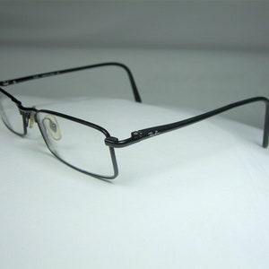 Puede incluir: Gafas de metal negro con montura fina y lentes transparentes. Las gafas tienen una montura rectangular fina y el logotipo de Ray-Ban es visible en el lateral.