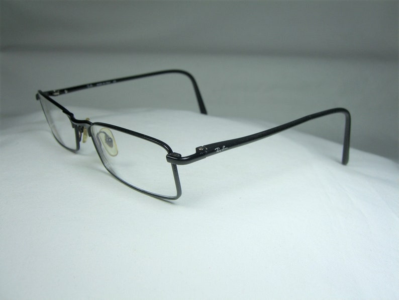 Puede incluir: Gafas de metal negro con montura fina y lentes transparentes. Las gafas tienen una montura rectangular fina y el logotipo de Ray-Ban es visible en el lateral.