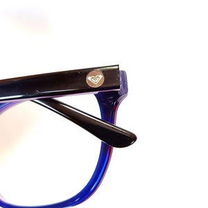 May include: Close-up of eyeglasses with a black frame and blue and red gradient arms. The glasses have a small logo on the top left corner of the frame. The glasses are set against a white background.
