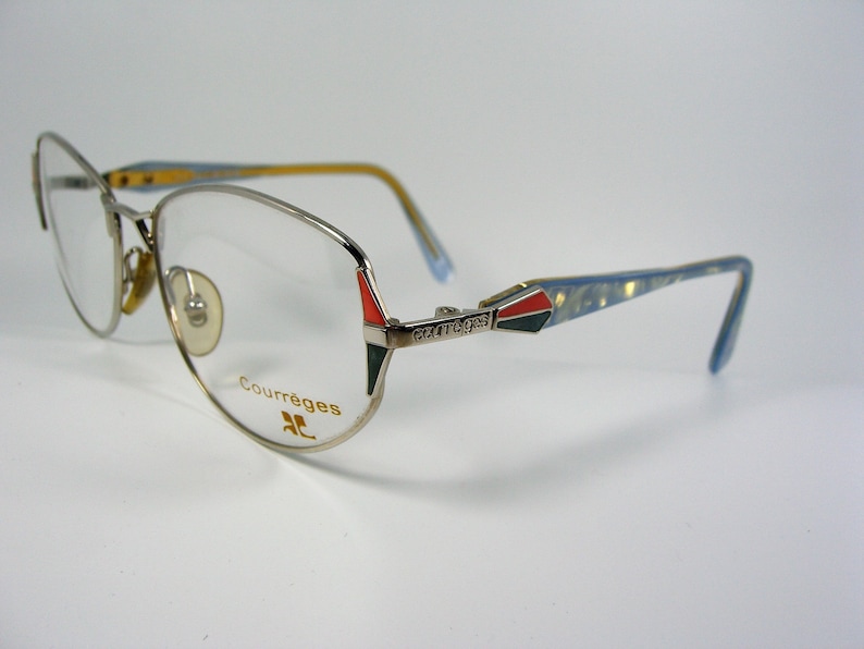 May include: Silver-toned eyeglasses with a blue and gold patterned arm. The frame has a red, black, and green accent at the temple. The brand name "Courr&egrave;ges" is printed in gold on the frame.