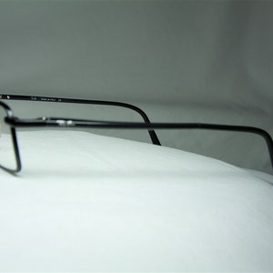 Puede incluir: Gafas de metal negro con un fino marco de metal y lentes transparentes. Las gafas est&aacute;n apoyadas sobre una superficie blanca.