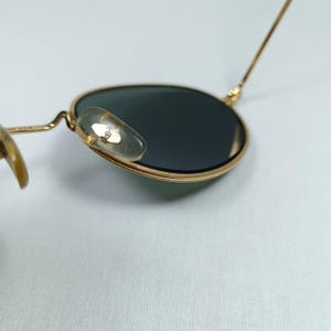 Puede incluir: Primer plano del marco de metal dorado de unas gafas de sol redondas. Las gafas tienen lentes verdes y un peque&ntilde;o detalle dorado en el lateral.