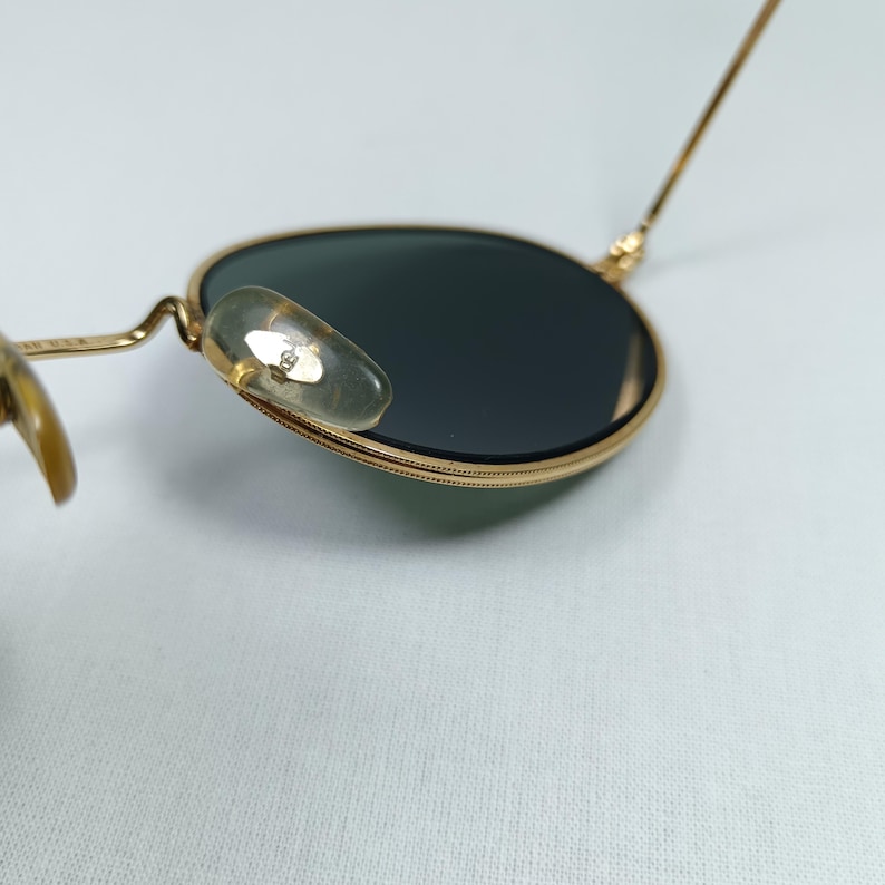 Puede incluir: Primer plano del marco de metal dorado de unas gafas de sol redondas. Las gafas tienen lentes verdes y un peque&ntilde;o detalle dorado en el lateral.