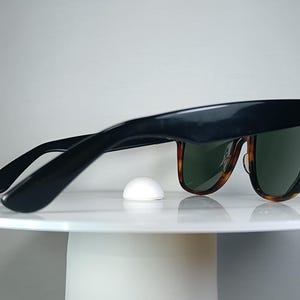 May include: Black sunglasses with a tortoise shell pattern on the lower frame and green lenses. The sunglasses have a gold accent on the temples and are resting on a white surface.