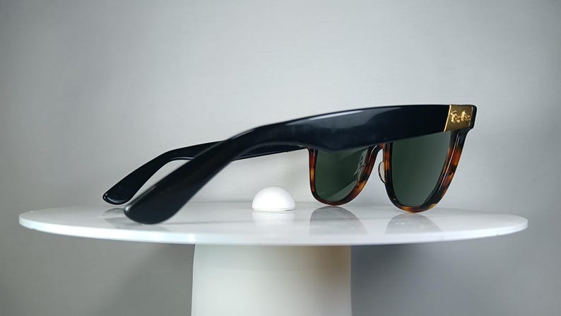 May include: Black sunglasses with a tortoise shell pattern on the lower frame and green lenses. The sunglasses have a gold accent on the temples and are resting on a white surface.